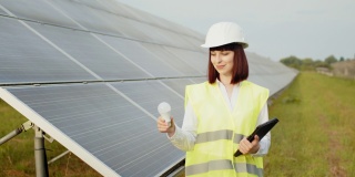 戴着头盔的女人站在太阳能农场拿着灯泡。一个穿着制服戴着白色头盔的女人站在太阳能农场。女工程师在工作中使用数码平板电脑。可再生电力的概念