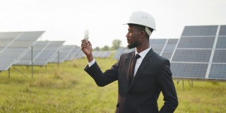 侧面的非裔美国商人在黑色的西装和白色的头盔拿着灯泡，而站在太阳能农场。生态和绿色能源的概念。非洲人拿着灯泡站在太阳能农场