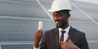 非裔美国人的肖像，戴着白色安全帽，穿着黑色西装，对着镜头微笑，手里拿着低能量灯泡。太阳能电池板站的背景。非洲人在太阳能农场拿着灯泡