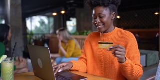 快乐的年轻女子拿着信用卡，在现代自助餐厅通过笔记本电脑在线购物