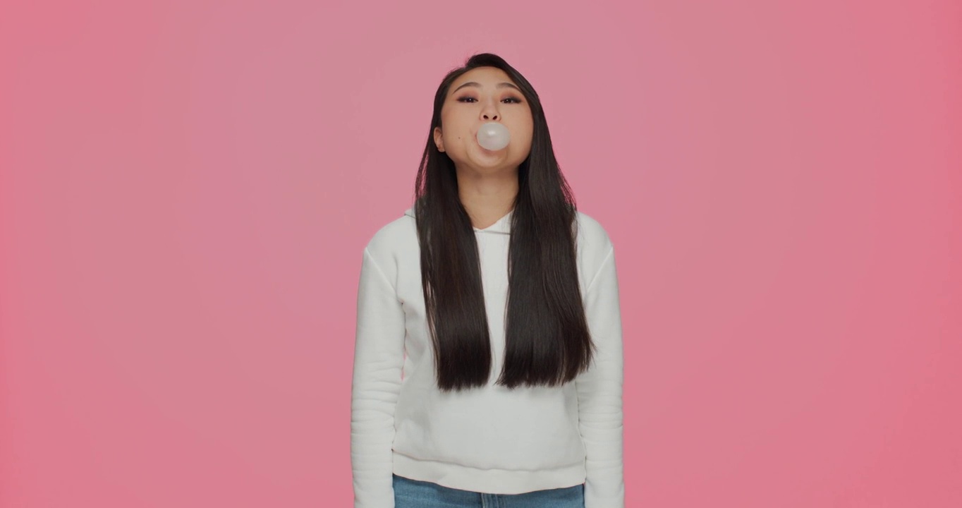 现代亚洲年轻女孩嚼泡泡糖，吹泡泡在粉红色的背景与广告copyspace。牙科保健和卫生