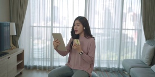 在家里工作的时候，一名亚洲女性在瑜伽球上放松、吃东西、在数码平板电脑上与同事们在线聊天。