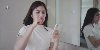 一位穿着睡衣的亚洲女性在早上在浴室刷牙准备上班时，通过智能手机查看电子邮件。