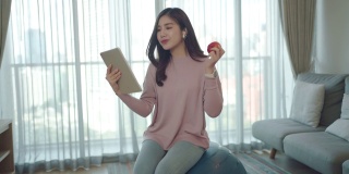 一名亚洲女性在家里工作的同时，在瑜伽球上放松，吃苹果，在数码平板电脑上与同事们在线聊天。
