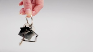 一个女性的手指握着和摇动金属钥匙链房子的新钥匙的特写镜头。灰色的背景。抵押的概念，购买房地产和租赁视频素材模板下载