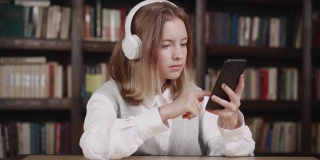 可爱的白人小女孩拿着智能手机玩手机应用，坐在图书馆里玩游戏。孩子在手机里学习，看视频，玩手机技术的概念