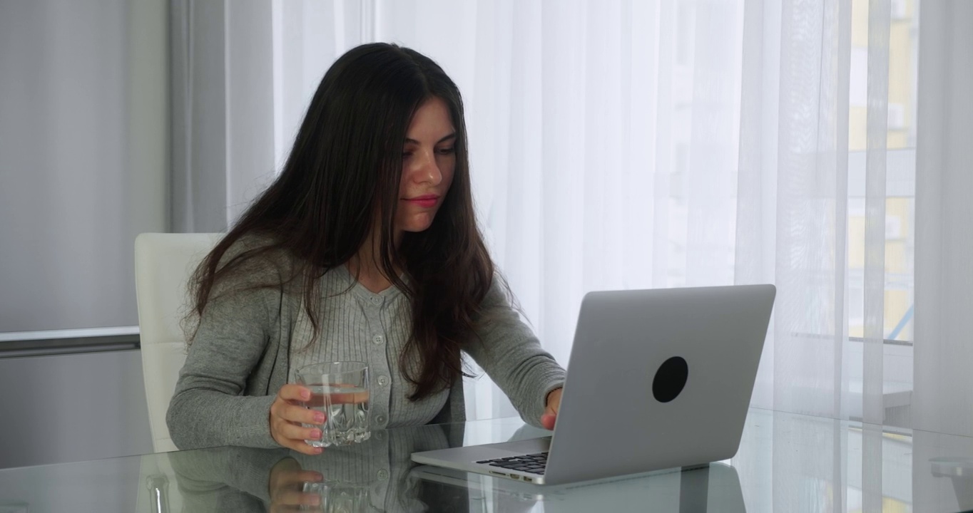 自由职业女性在家里用笔记本电脑工作。一个穿着灰色毛衣的白人女孩坐在家里厨房的桌子旁喝着水，开始打字。特写镜头