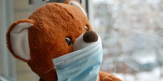 在新冠肺炎隔离期间，戴着防护口罩的好奇泰迪熊透过窗户看着外面的雪花