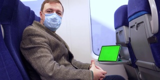 穿着外套、戴着医用口罩的千禧一代男子坐在火车上，看着摄像机，他面前的折叠桌上放着一台带有色度键的绿色屏幕的平板电脑