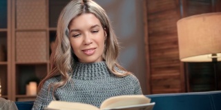 微笑的年轻金发女性享受阅读有趣的书翻页在舒适的家