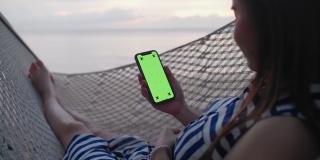 一个年轻的女孩躺在沙滩上的吊床上使用智能手机