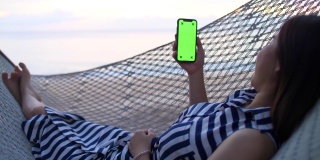 一个年轻的女孩躺在沙滩上的吊床上使用智能手机