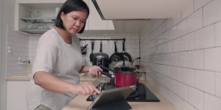 亚洲妇女在厨房做饭，同时看平板电脑的食谱