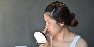 忧虑的亚洲妇女与坏情绪，而看着痤疮和疤痕出现在她的脸上的迷你镜子。