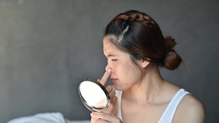 忧虑的亚洲妇女与坏情绪，而看着痤疮和疤痕出现在她的脸上的迷你镜子。视频素材模板下载