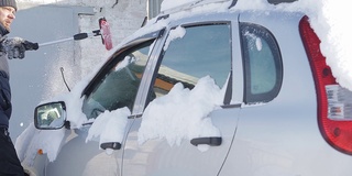 在阳光明媚的冬日，一名男子正在清理汽车上的积雪，这是反常的降雪和气候变化