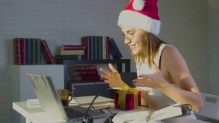 圣诞前夜，一名戴着圣诞帽的年轻女性在办公室里收到了一份礼物，她通过在线视频电话感谢了自己的丈夫视频素材模板下载