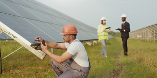 能源技术员戴着头盔，穿着制服，在户外用万用表测量太阳能电池板的电压。一群工程师用平板电脑和剪贴板站在背景上。靠近太阳能电池板的男子