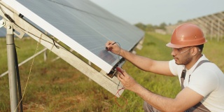 有能力的技术员戴着安全帽和眼镜，用万用表测量太阳能电池板的安培数。印度男子在户外修理光伏电池。绿色能源的概念。太阳能电池板的安培数