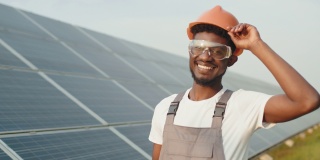 非裔美国人，戴着安全眼镜和头盔，拿着剪贴板，站在太阳能板上。概念服务工作和替代能源。检查太阳能电池板的技术员