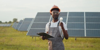 非洲裔美国人站在场地上，手里拿着太阳能电池板和灯泡。男性技术员穿着棕色工作服，戴着橙色头盔和安全眼镜。关于绿色能源的想法。太阳能农场