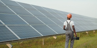 能源工程师带着仪器在太阳能农场上行走。非洲裔美国人在制服和头盔使用仪器，而工作的领域与太阳能电池板的后视图。生产能源