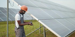 非洲专业工程师戴着头盔，穿着制服，用万用表测量太阳能电池板的安培数。人、维护和替代能源的概念。穿着工作服的人在测量阻力