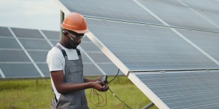 身穿灰色工作服的非裔美国人在户外测量太阳能电池板的阻力。穿着工作服的非洲人在测量太阳能电池板的阻力。工程师测量太阳能电池板的安培数