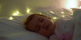 1-2岁的小女孩穿着粉红色的睡衣睡在白色的床上。孩子晚上有放松的休息时间枕头上的花环。童年、圣诞节、亲子观念