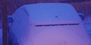 在冬天，黄昏时分，停在停车场路灯下的汽车被雪覆盖。城市街道上大雪纷飞。