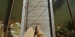 慢镜头拍摄的一名潮人走在一座木桥上，桥横跨山间河流。男人的腿在休闲旅行靴子的俯视图