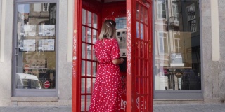 一个穿红衣服的年轻女子走近一个红色的电话亭，使用城市电话