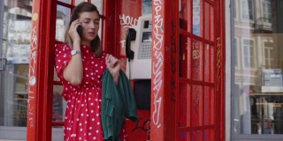 一个年轻女子站在一个旧的红色电话亭里用手机聊天