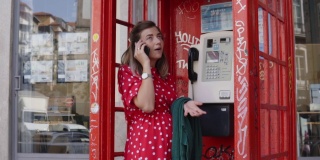 一个年轻的女人站在红色的旧电话亭里用她的小玩意说话