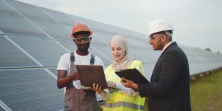 戴头巾的穆斯林妇女和戴白帽的印度男子与太阳能站的非裔美国技术人员交谈。多种族的人使用笔记本电脑，剪贴板和蓝图在户外工作