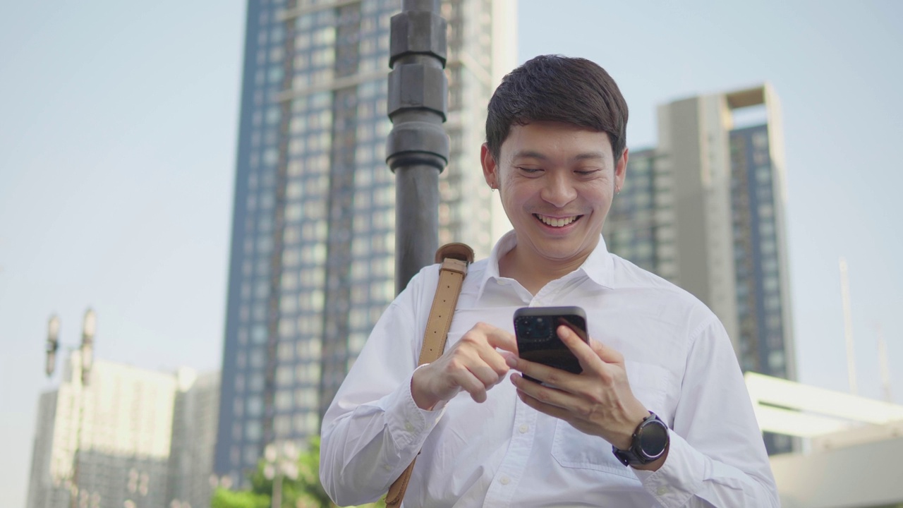 一位面带微笑的亚洲商人站在城市外的街道上，用智能手机上网。