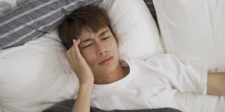 高角度的观点与头部射击不高兴的亚洲人躺在床上醒着揉他的太阳穴与皱眉，而遭受严重的头痛早上在家里。