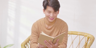 快乐英俊的亚洲男人坐在沙发上微笑和阅读书籍放松时间在家里。兴高采烈的商人穿着蓝色衬衫，坐在沙发上看书