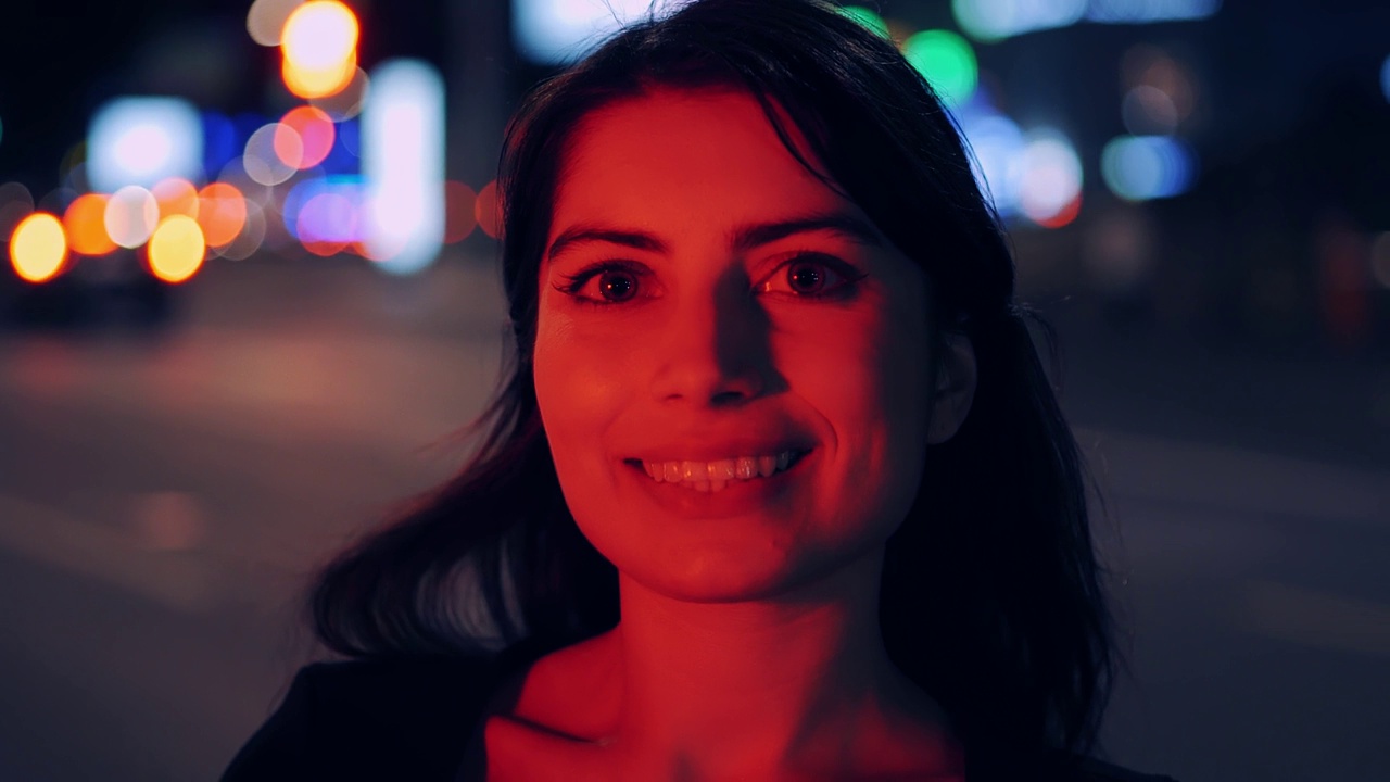 美丽的黑发女人微笑的肖像，看着相机。夜城街道背景散景