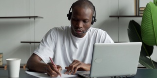 专注的黑人商人戴着耳机，在笔记本上写笔记，观看视频课程网络研讨会