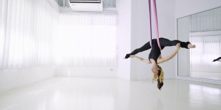 苗条运动年轻女子在黑色运动服展示瑜伽飞行在吊床上。一名年轻的亚洲女子在白色的体操室内进行反重力空中练习，手拿粉色丝带倒挂在空中。