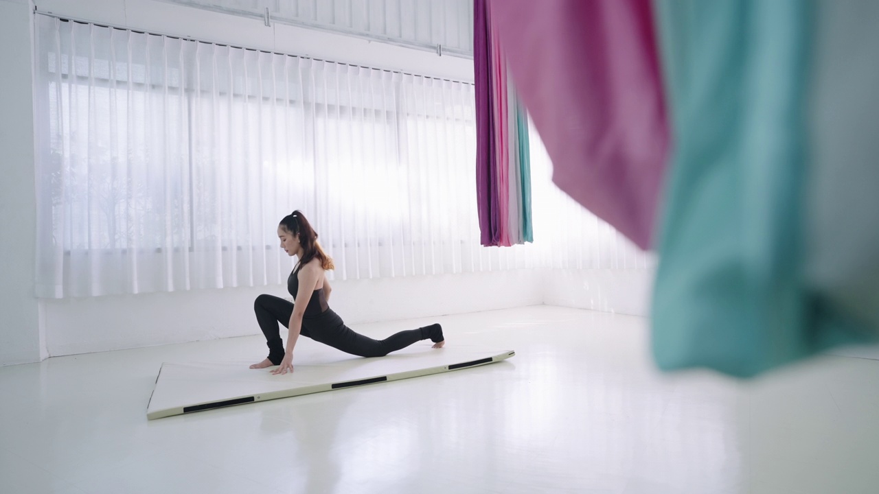 年轻的亚洲妇女训练瑜伽姿势新月形弓步在膝盖上，手放在床垫上。年轻苗条的女孩穿着黑色运动服，在健身室的白色床垫上做有氧伸展运动，舒缓肌肉紧张，增强柔韧性