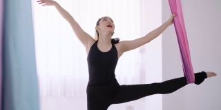 年轻漂亮苗条的身体健身亚洲妇女站在一只脚和伸展她的腿与彩色丝带。年轻迷人的熟练女子在黑色的紧身衣锻炼做伸展瑜伽姿势使用吊床在健身房。