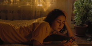 一个漂亮的年轻女人正躺在床上，靠在她的胳膊上，用着智能手机。窗外的晚霞，傍晚