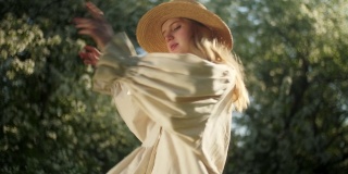 富有表现力的年轻金发女人在强烈的阳光下穿着白色连衣裙和帽子的肖像，快乐地跳舞在模糊的开花树背景与自然散景在柔和温暖的日落背光户外。慢动作