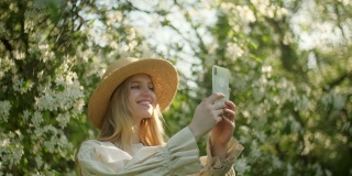 年轻美丽的金发女子在春天的花圃里用智能手机拍照的肖像。春天公园里快乐美丽的女孩。花开在树上。在花树旁摆姿势的女人