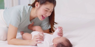 新亚洲妈妈玩可爱的新生婴儿在床上微笑和幸福在家里。妈妈和婴儿说话，牵着她的手一起笑。婴儿和母亲节的概念