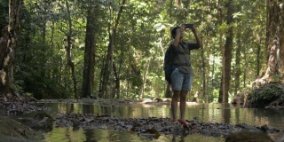 夏季，亚洲女性徒步旅行者背着背包，站在热带森林的小溪中，用手机拍照。