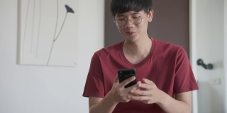 亚洲男子戴着眼镜坐在沙发上的早上，他的手机，收到短信，非常高兴和微笑。4 k决议。