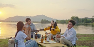 小组四人朋友亚洲男女野营，喝啤酒，庆祝，唱歌，弹吉他，玩乐，享受地面帐篷。日落假期期间的水库区域。